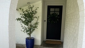 A white brick exterior with a concrete pathway leading to a dark door. A large blue pot with a tree is placed on the left.