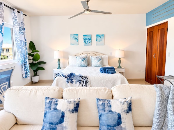 A bedroom with a bed, two bedside tables, a ceiling fan, a window with curtains, and a view of buildings outside.