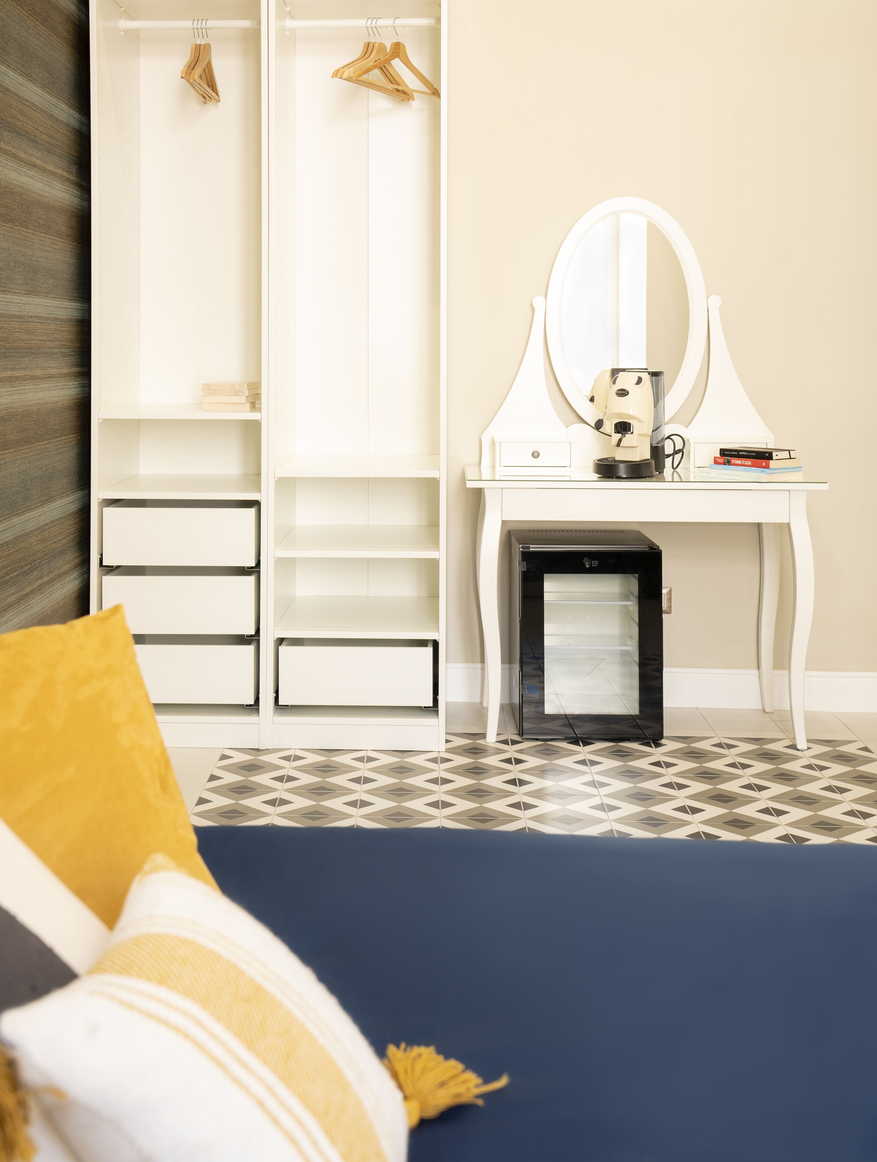 A bedroom with a bed, a white vanity with a mirror, a small heater, and a wardrobe with hanging clothes.