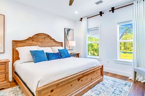 A spacious bedroom with a large wooden bed, a nightstand, a bedside table, and a window with a view of greenery.