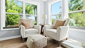 Una habitación con dos sillones tapizados, una mesita de noche con una lámpara y un reposapiés tejido.