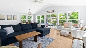 Un salón espacioso con un sofá seccional azul oscuro, una mesa de centro de madera y un sillón a rayas. Grandes ventanales ofrecen vistas al exterior.