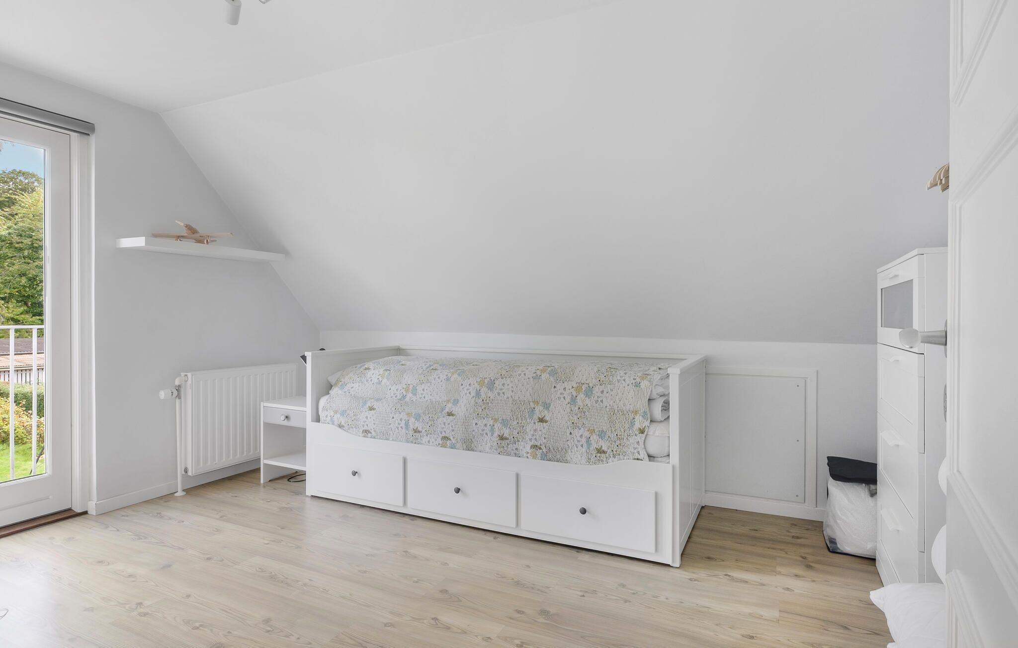 A bedroom with a sloped ceiling, a white bed with a floral duvet, a white dresser, and a radiator.