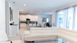 Un salon moderne avec une table basse en verre, un canapé blanc et un coin repas avec une cuisine en arrière-plan.