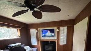 Een gezellige woonkamer met een donkergrijze bank, een plafondventilator en een televisie waarop een scène van de politie te zien is.