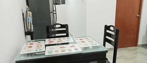 A dining table with four chairs, a refrigerator, and a door in a room with white walls and a tiled floor.