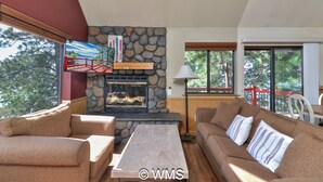 A cozy living room with a stone fireplace, comfortable seating, and large windows offering a view of trees.