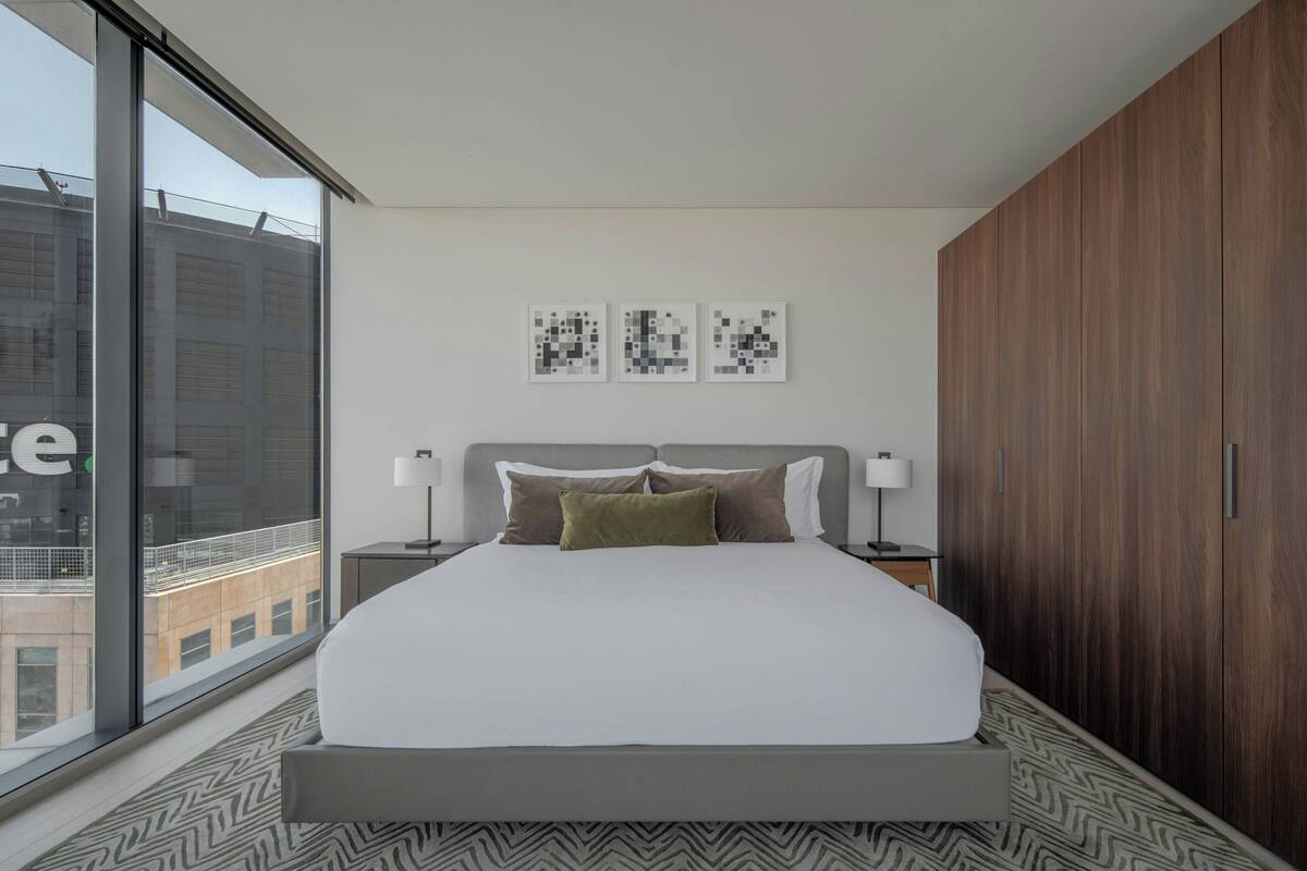 A modern hotel room with a large bed, two bedside tables, a window with a view of buildings, and a wooden wardrobe.