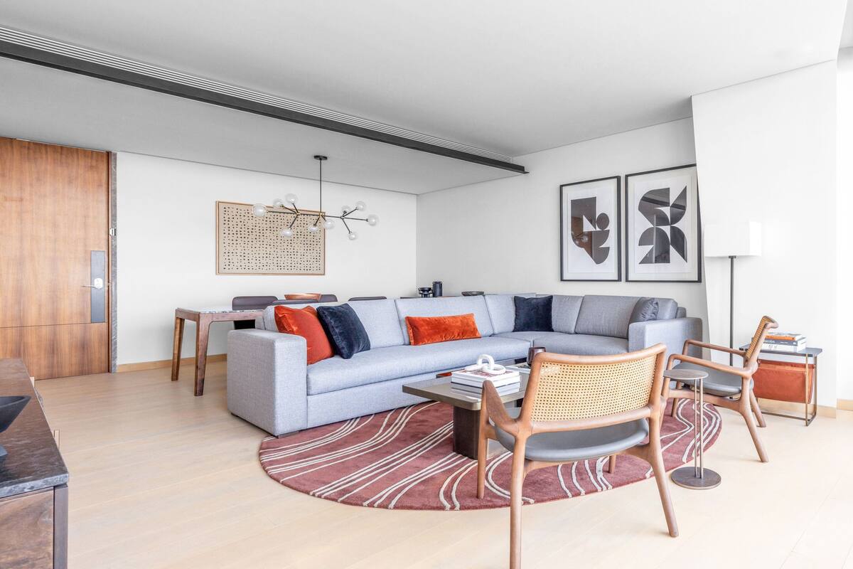 A modern living room with a sectional sofa, a coffee table, and two chairs. There is a dining table with chairs, a lamp, and artwork on the wall.