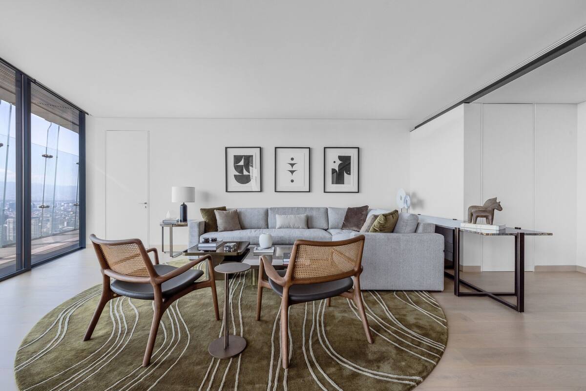 A modern living room with a sofa, two chairs, a coffee table, and a small table with a lamp. There are three framed artworks on the wall.