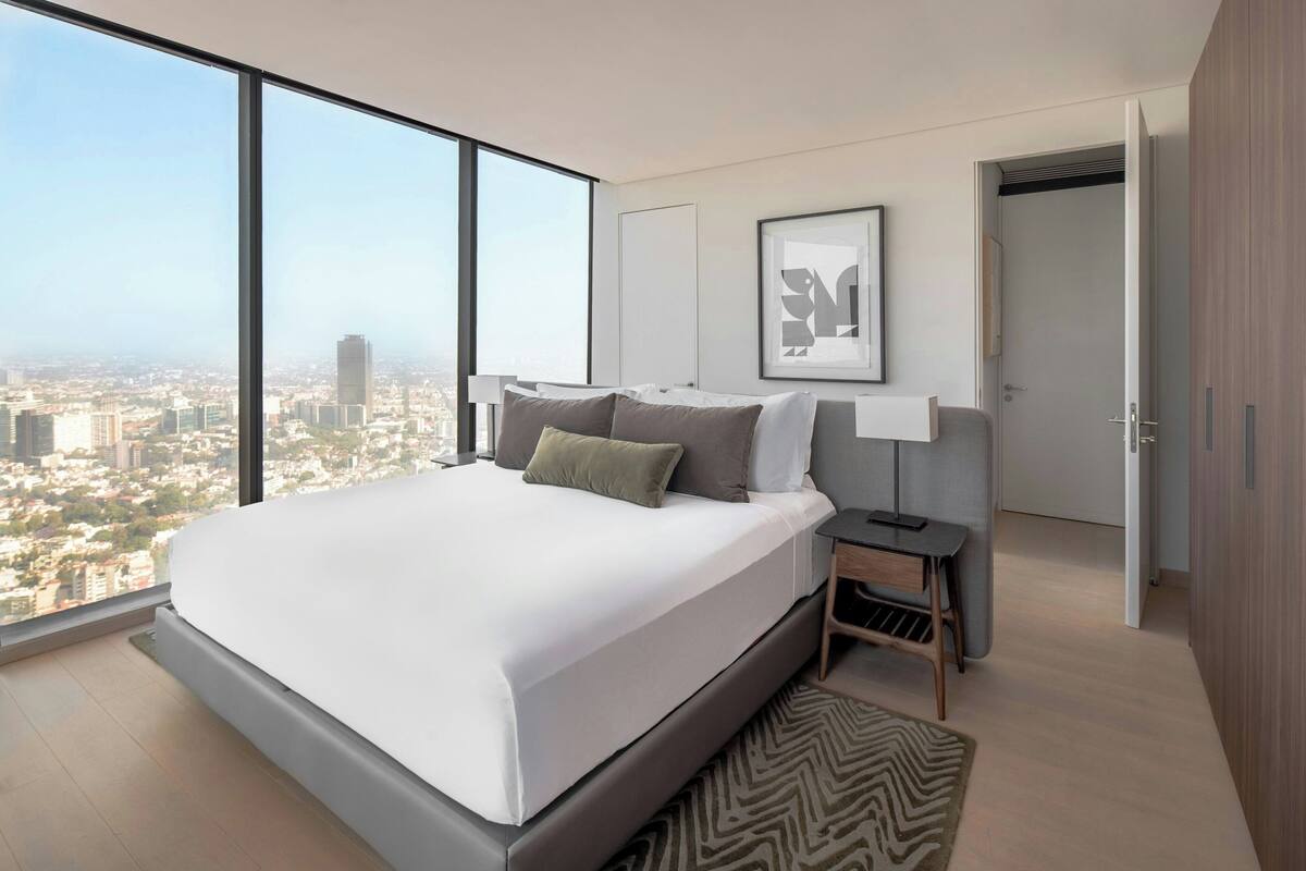 A modern bedroom with a large bed, a nightstand, a lamp, and a view of the city.