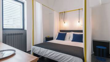 Un dormitorio con una cama con dosel, una mesita de noche, una ventana con persianas y una pequeña mesa redonda.