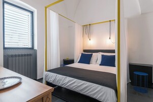 Un dormitorio con una cama con dosel, una mesita de noche, una ventana con persianas y una pequeña mesa redonda.
