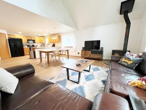 Een moderne woonkamer met een bruine leren bank, een salontafel en een televisie. Achtergrond is een keuken met houten kasten en een zwarte koelkast.