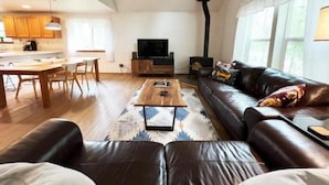 A modern living room with a sofa, coffee table, and a TV. There is a kitchen area with wooden cabinets and a dining table.