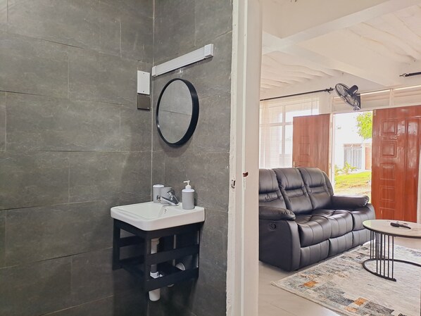 A modern bathroom with a grey tiled wall, a round mirror, a white sink, and a black stool. - Cave Diani Studio Apartments (Diani Beach)