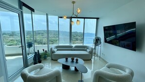 Ein modernes Wohnzimmer mit einer Couch, zwei Sesseln, einem Couchtisch und einem Fernseher. Große Fenster bieten Blick auf Grünflächen und Hügel.