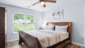 Een slaapkamer met een bed, een plafondventilator, een raam met jaloezieën en een nachtkastje met een lamp.