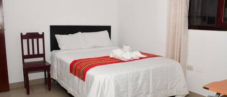 Una cama bien hecha con colcha blanca y manta a rayas dorada y roja, una silla de madera y una ventana con cortinas color crema.