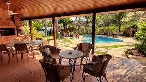 A covered patio with outdoor seating, a brick fireplace, and a pool area with lounge chairs. - Astral Nature (Pedra Verde (Domingas Dias) Ubatuba)