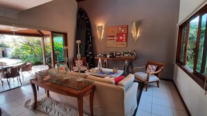A spacious living area with a glass coffee table, beige sofas, and a dining table with chairs. There is a bar area with bottles and glasses, and a large window offering a view of a pool and greenery. - Astral Nature (Pedra Verde (Domingas Dias) Ubatuba)