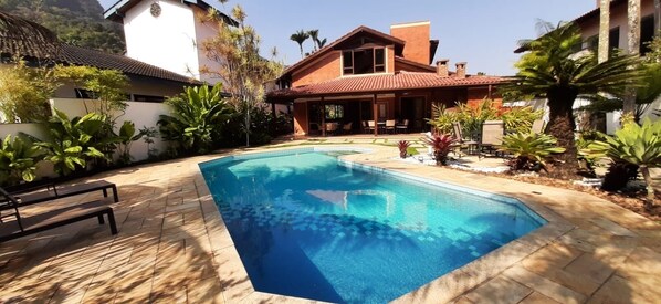 A swimming pool with a tiled deck, surrounded by various plants and a building with a tiled roof.