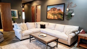 Una sala moderna con un sofá seccional color beige, una mesa de centro de madera y un cuadro grande con un paisaje.