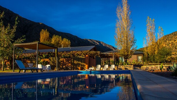 Un área de piscina con sillas de descanso y una pérgola, rodeada de cactus y árboles, con montañas al fondo.
