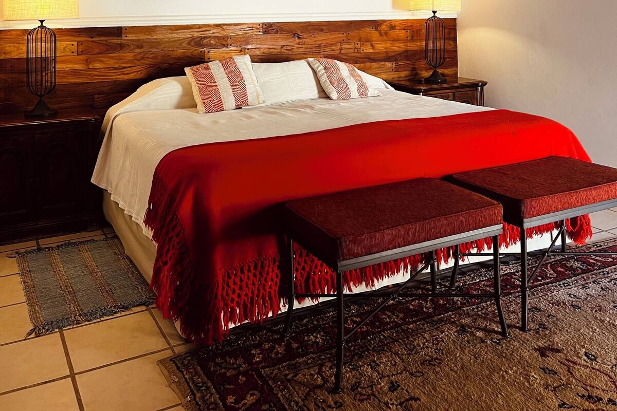 Un dormitorio con una cama grande, cabecera de madera, una manta roja, un banco, un cuadro en la pared y una araña.
