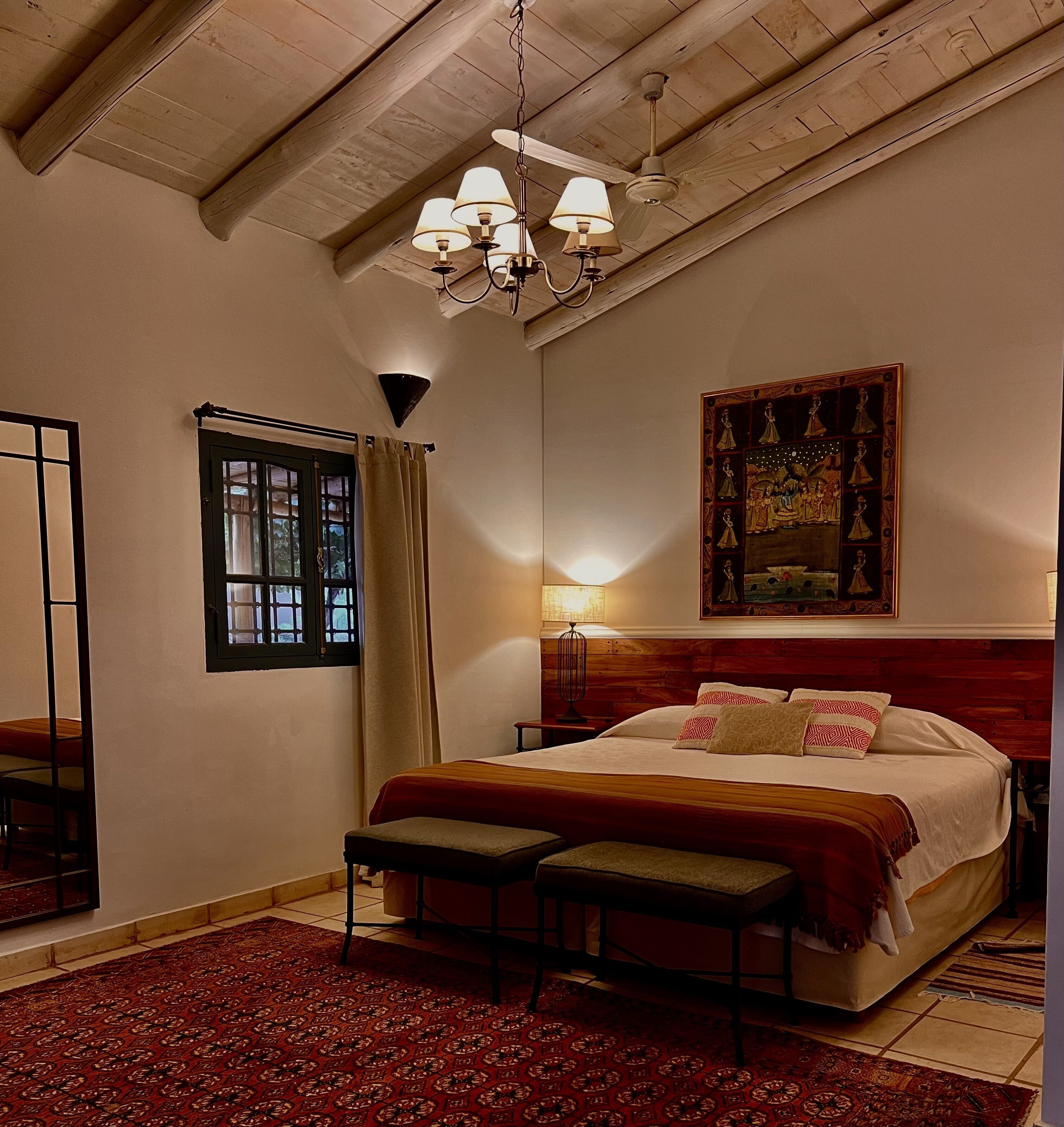 A bedroom with a large bed, a bench, a window with curtains, a chandelier, and a painting on the wall.