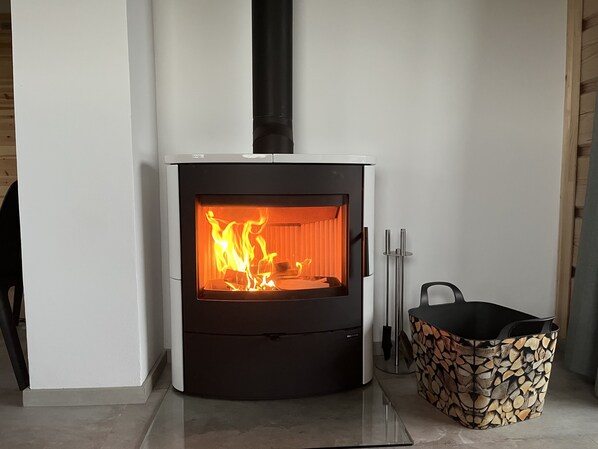 A modern wood-burning stove with a glass front, a black chimney, and a log basket beside it. - Moderne Blockhütte im Erzgebirge - Roubenky Hrebecna 212 (Karlovarský kraj)