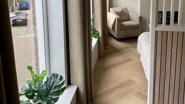 A narrow hallway with a window, a radiator, a potted plant, a bed, a chair, and a wall with two framed pictures.