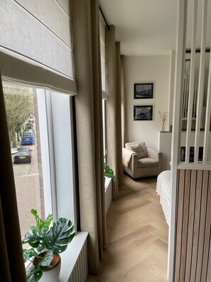 A narrow hallway with a window, a radiator, a potted plant, a bed, a chair, and a wall with two framed pictures.