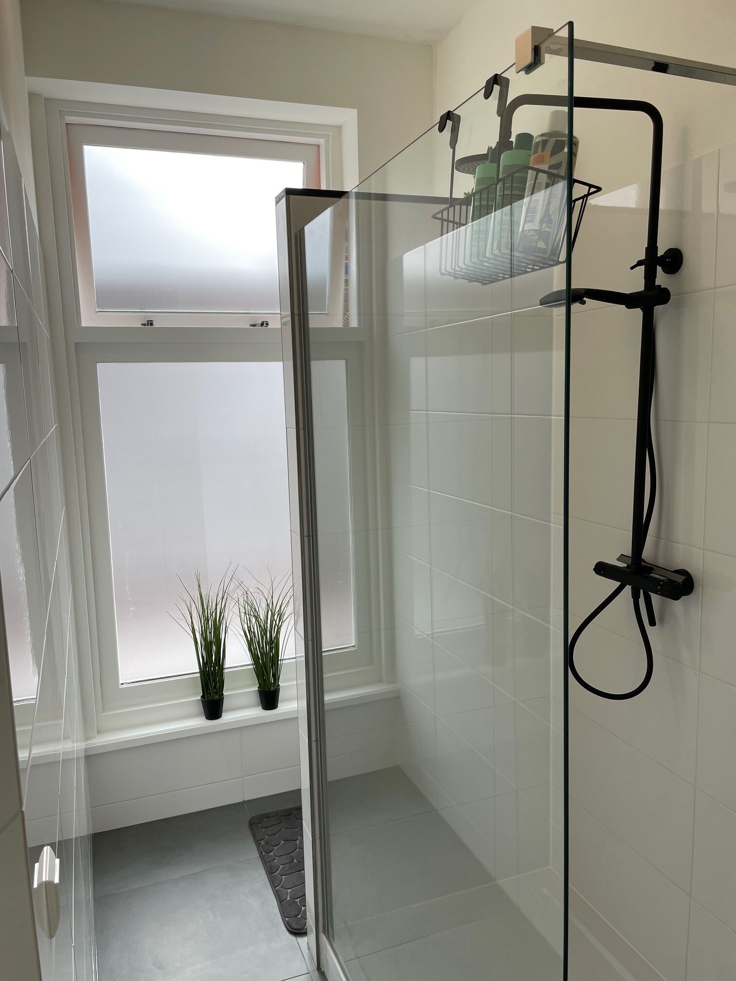Un baño moderno con cabina de ducha de vidrio, paredes con azulejos blancos, un ventanal con vidrio esmerilado y una pequeña planta en maceta sobre el alféizar.
