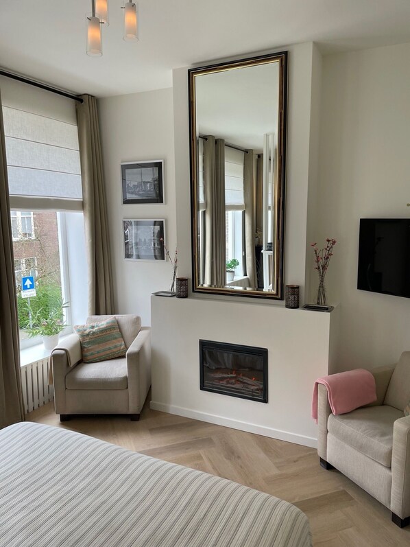 A modern hotel room with a large bed, a fireplace, a TV, and a large window with curtains.