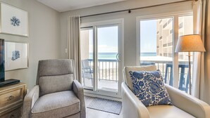 Ein Zimmer mit einer Couch, einem Sessel und einem Fernseher auf einer Holzkommode. Es gibt ein großes Fenster mit Blick auf den Strand und einen Balkon.