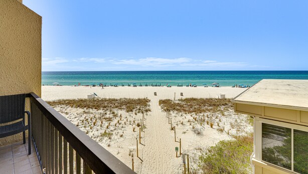 Blick auf den Strand vom Balkon aus, mit einem Sandweg, der zum Wasser führt, einem Strandsonnenschirm und ein paar Personen in der Ferne.