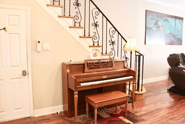 A piano in a room with a wooden floor, a staircase with black railings, a lamp, and a painting on the wall.