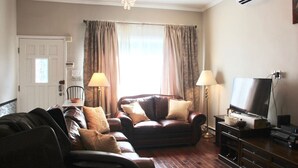 A living room with a brown leather sofa, two lamps, a television on a wooden stand, and a window with curtains.