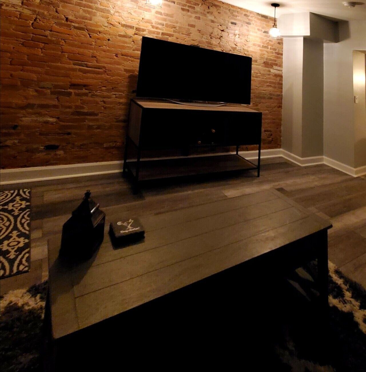 A room with a brick wall, a flat-screen TV mounted on a stand, a dark wooden coffee table, and a patterned rug.