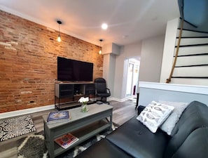 A modern living room with a brick wall, a flat-screen TV, a black leather sofa, a coffee table, and a staircase. - Historic 2BR Fishtown Rowhome with Private Patio & Prime Location (Philadelphia)