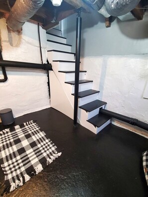 A staircase with black railings leading to a lower level, a black and white patterned rug on the floor, and a cylindrical object on the left.