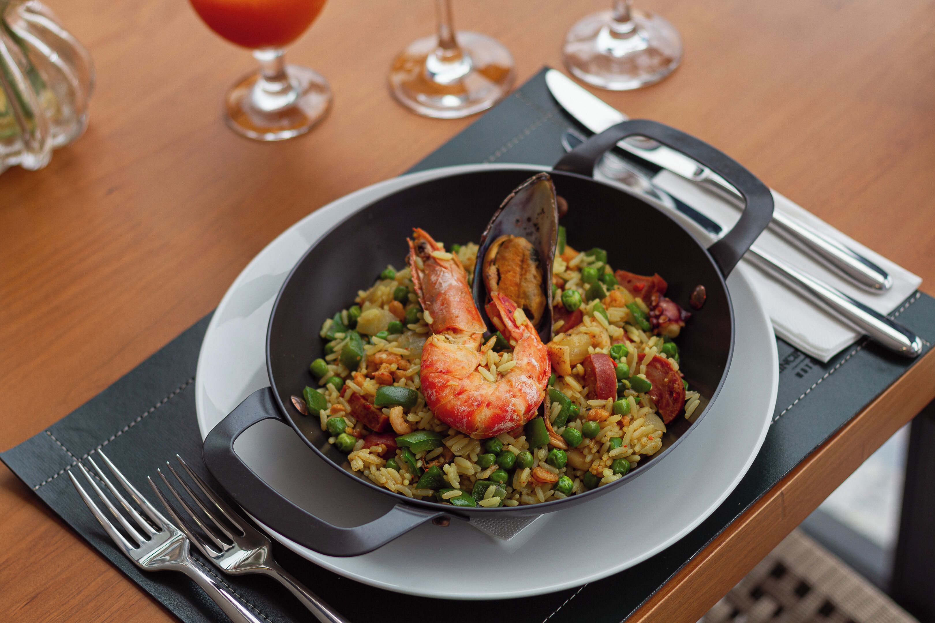 Une portion de paella aux fruits de mer servie dans un plat, accompagnée d’une fourchette et d’un couteau posés sur une serviette, avec des verres à vin en arrière-plan.