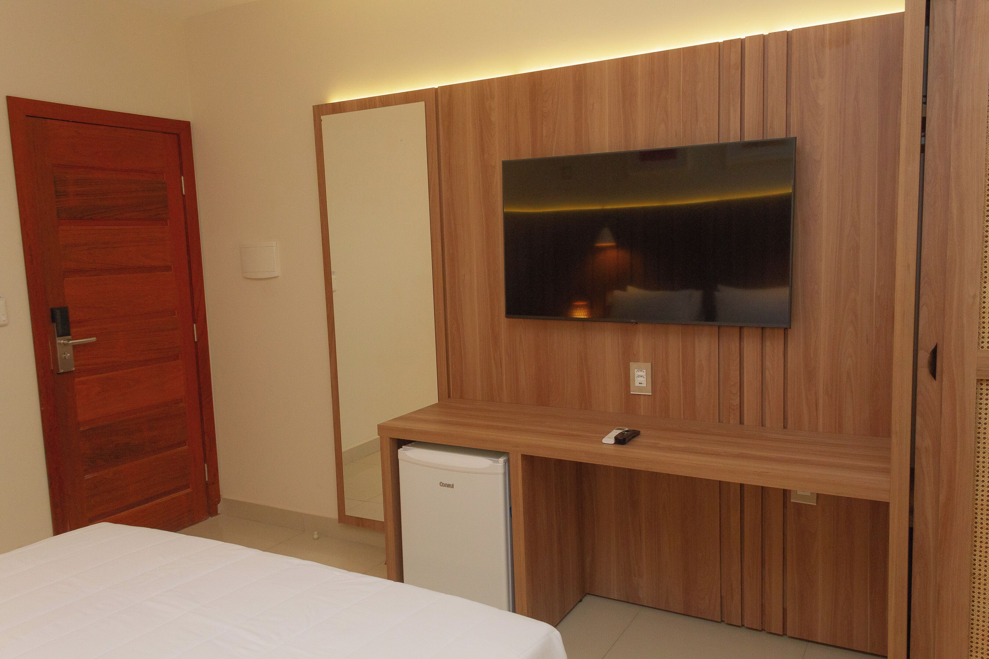 A hotel room with a wooden panel wall, a television mounted above a desk, a small refrigerator, and a bed with a white bedspread.