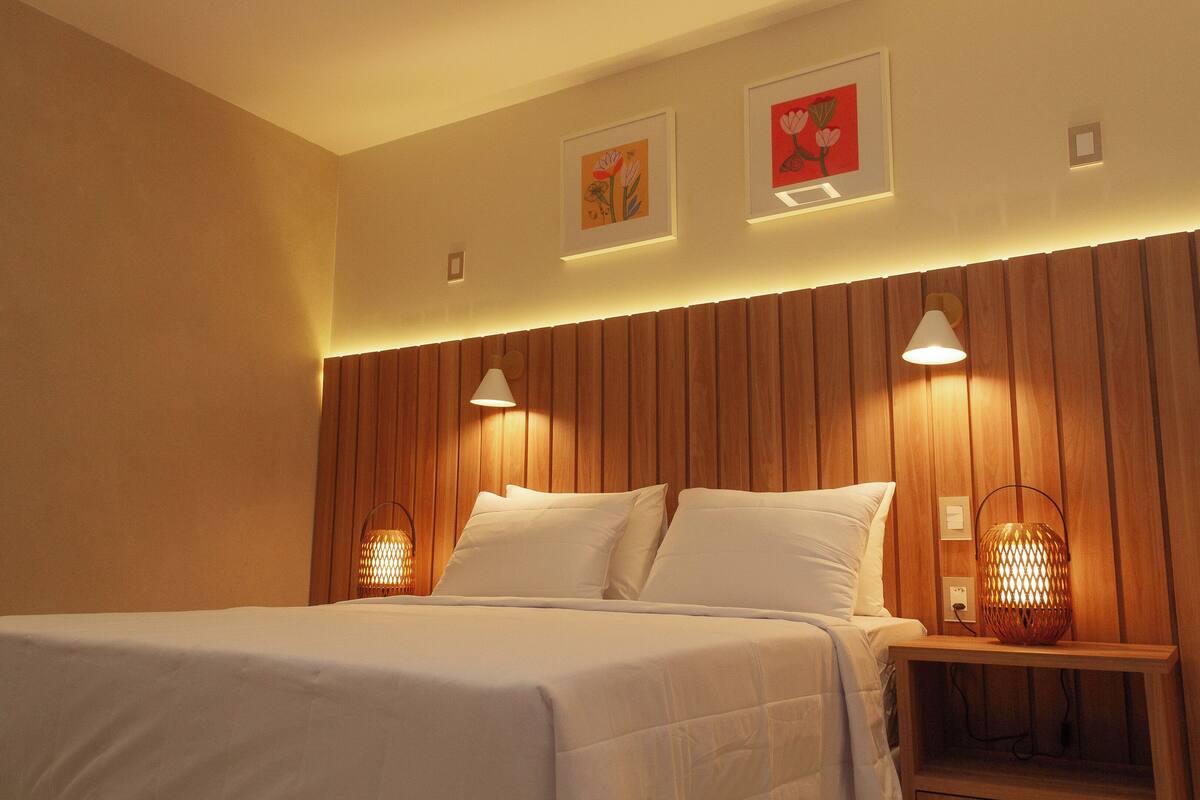 Una cama bien hecha con sábanas blancas, dos lámparas fijadas a la pared y dos cuadros florales enmarcados.