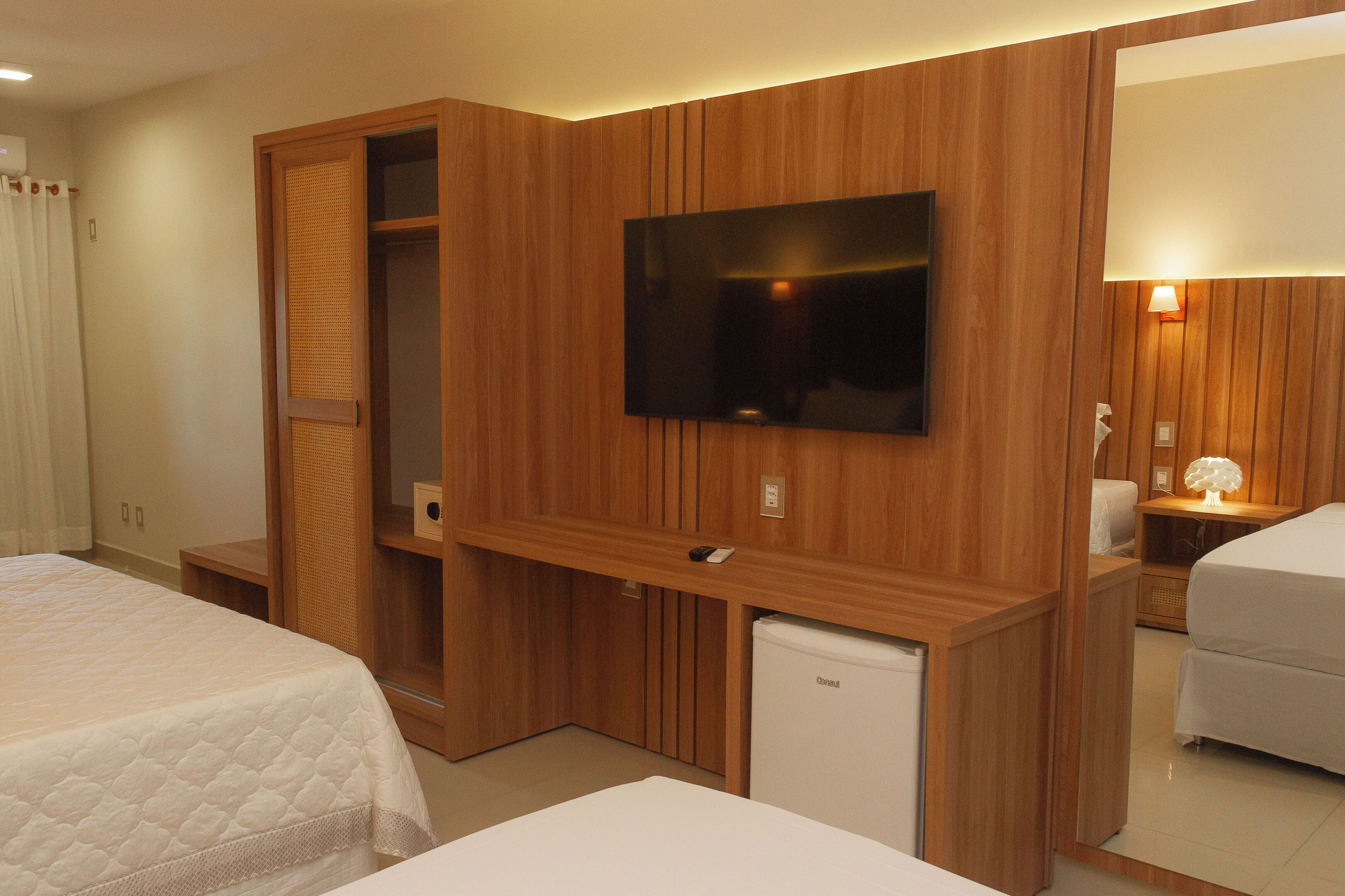 Una habitación de hotel con cama, televisión montada en la pared, un refrigerador pequeño y un armario de madera.