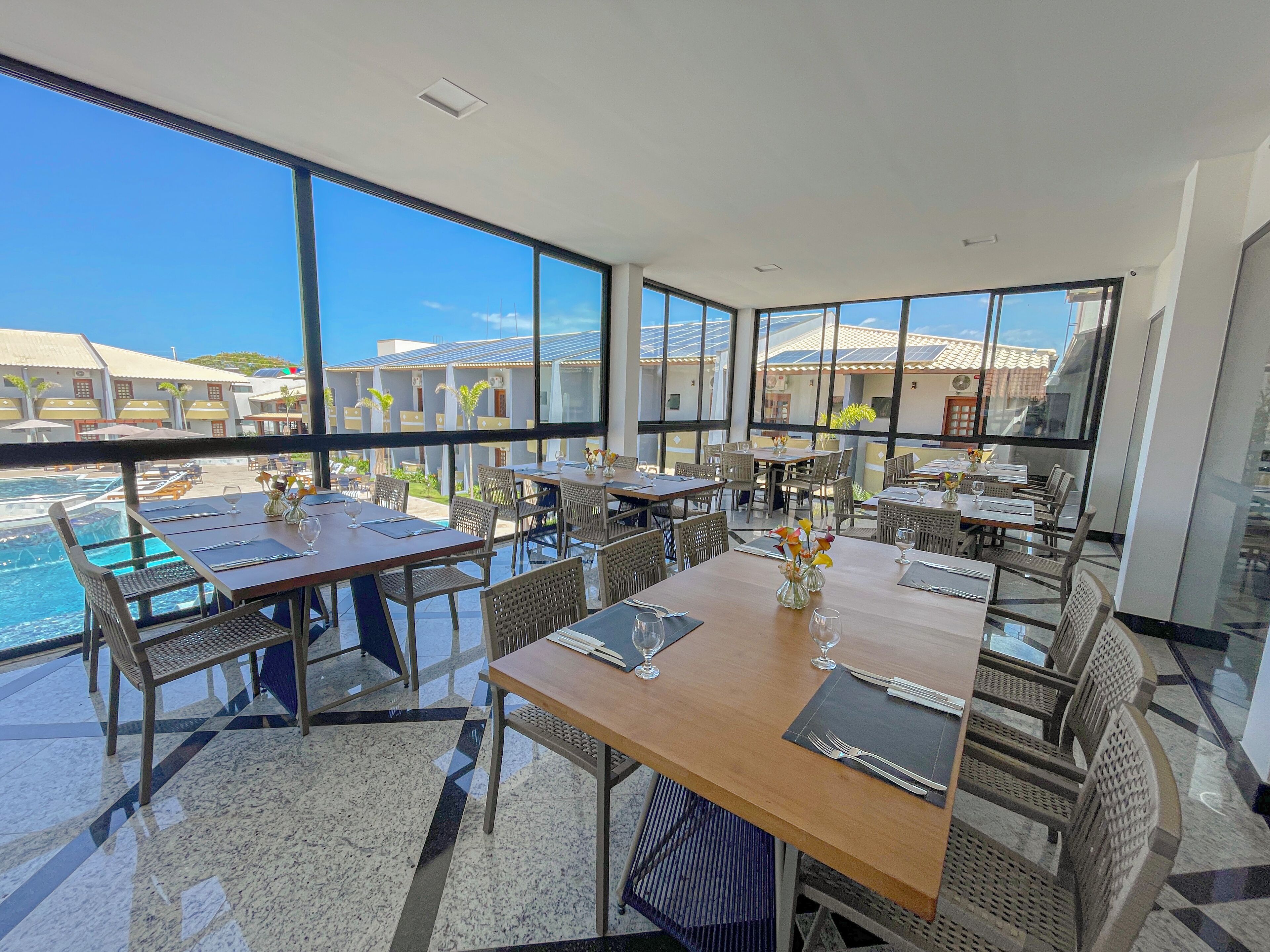 Un comedor amplio con ventanas grandes que ofrecen vista a una piscina y edificios. Las mesas están dispuestas con sillas y centros de mesa.