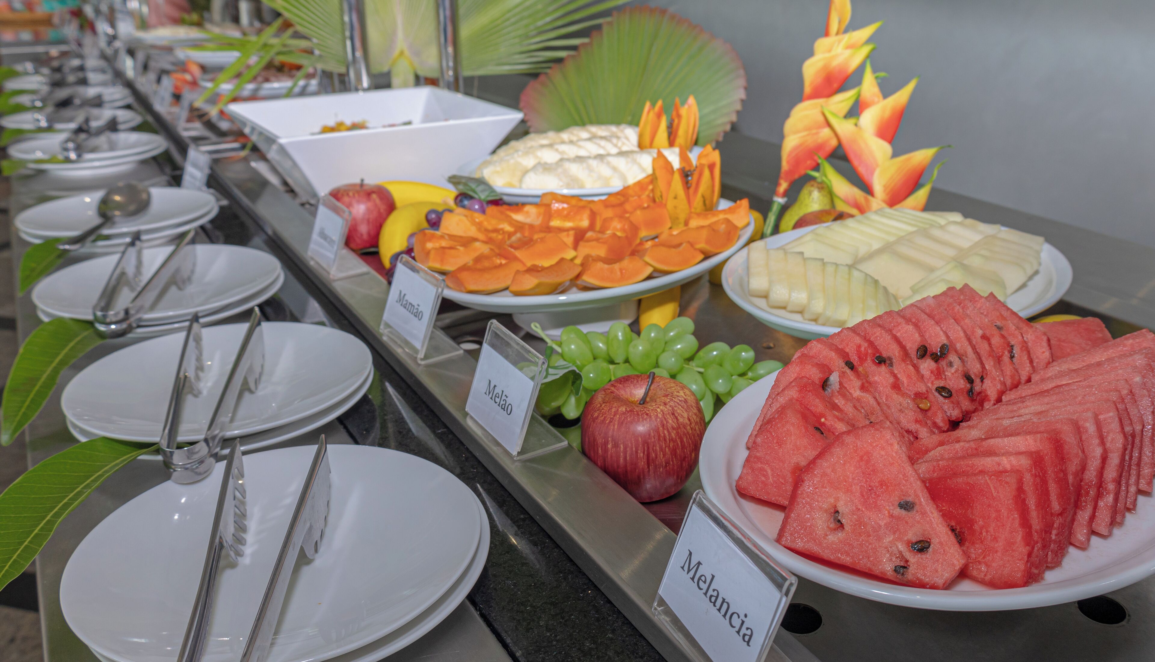 Buffet com diversas frutas e legumes, incluindo melancia, uvas e maçãs, dispostos em pratos e tigelas.