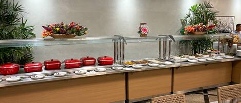 A buffet area with a variety of food items, a glass display case, and a seating area with wooden chairs and a table set for dining.