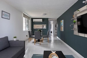 Una sala de estar moderna con un sofá gris, comedor con sillas, televisor montado en la pared y una cocina al fondo.